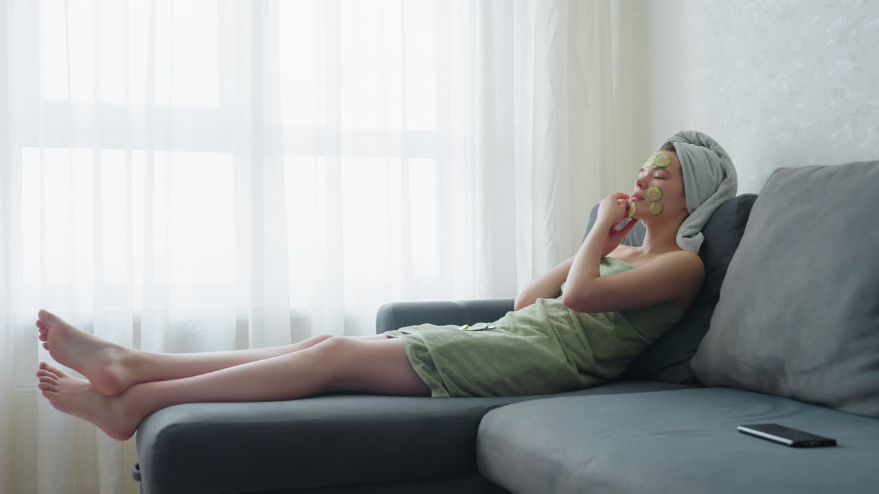Woman wrapped in towel, relaxing on couch next to window, placing cucumber slices on her face, enjoying calming self-care routine for rejuvenating facial treatment and skin relaxation