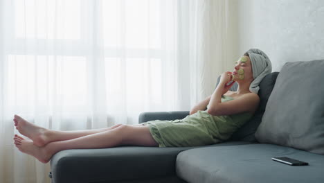 woman wrapped in towel, relaxing on couch next to window, placing cucumber slices on her face, enjoying calming self-care routine for rejuvenating facial treatment and skin relaxation