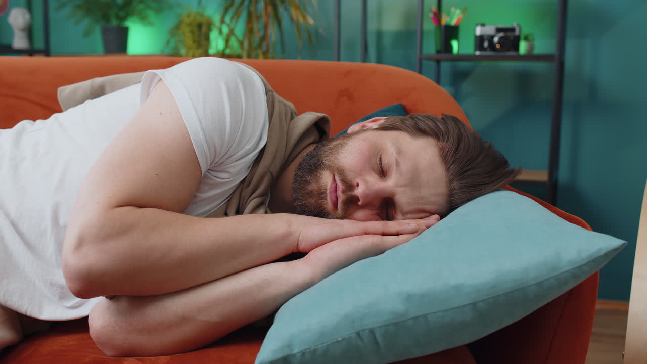 Tired young man lying down on comfortable couch taking a rest daytime nap relaxing falling asleep