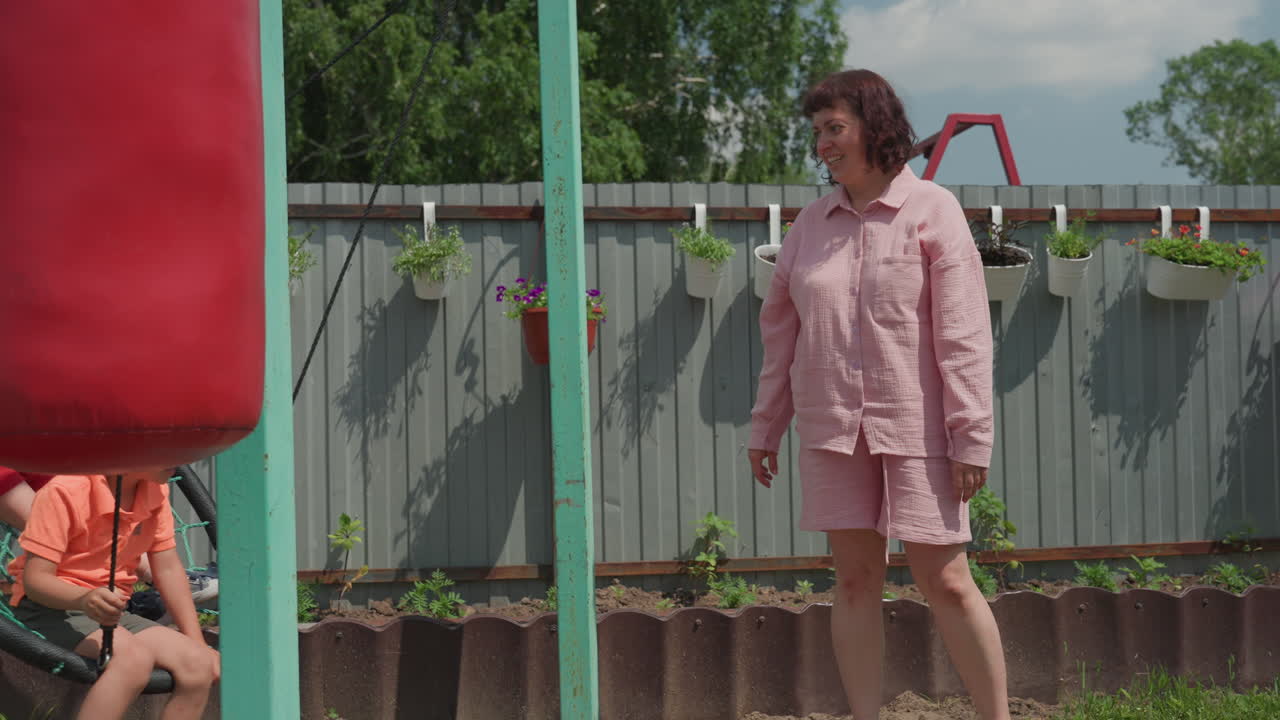 Caucasian Woman In Pink Coat Guiding Boys On Nest Swing In Backyard, Offering Gentle Push And Warm Smile, Summer Afternoon Near Fence And Garden, Nurturing Scene Of Parenting And Play