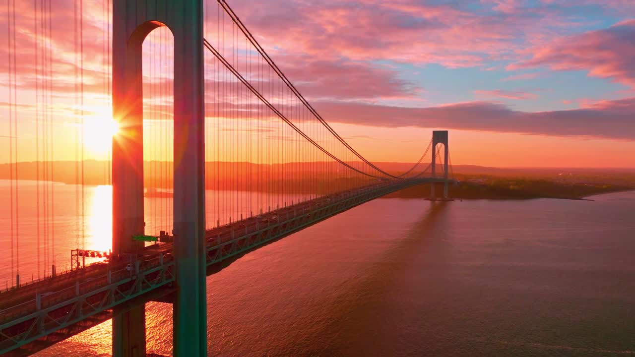 Veduta aerea con drone del ponte Bronx-Whitestone a New York City all'ora d'oro, con traffico che attraversa il ponte sospeso sopra acqua calma e un cielo radioso all'alba.