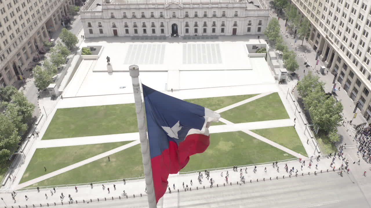 Aerial enormes flag with government palace in the back with people walking and surrounded by buildings in Santiago de Chile
