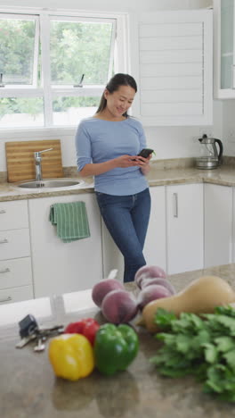 video vertical de una mujer asiática sonriente de pie en la cocina usando un teléfono inteligente