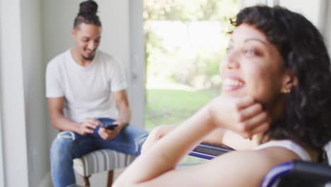 mujer biracial sonriente en silla de ruedas en casa con un compañero masculino usando un teléfono inteligente