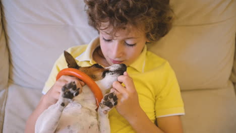 Blond-Boy-With-Curly-Hair-Sitting-On-The-Couch-Caresses-His-Dog