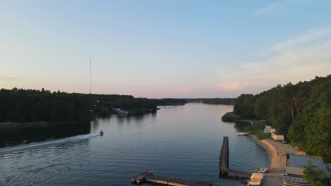 a drone captures a lakeside dock at twilight as a jet ski skims calm water, creating gentle ripples under a pastel sky framed by a forested shoreline.