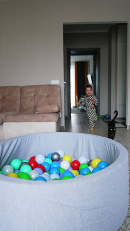 a dry pool with colorful balls is in the living room. toddler boy runs into the room and dives into the pool. black cat runs with a kid. vertical video.