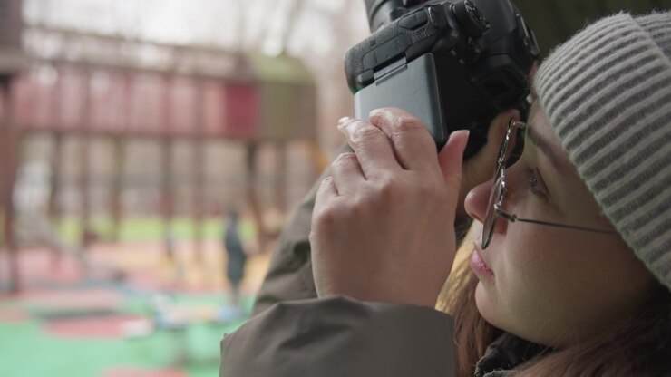 Asian woman adjusting DSLR settings, checking viewfinder and turning dials, testing exposure and focus, closeup hands on camera, patient concentration and careful technique in park
