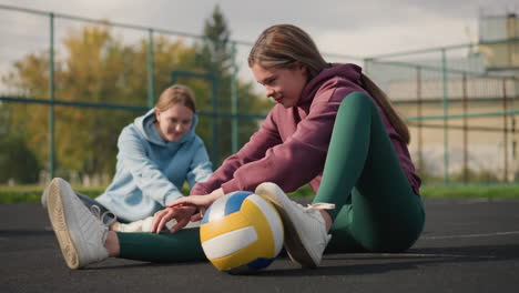 ladies working out seated on the ground with volleyball placed under one leg, outdoor court setting with iron fence, trees, and a building in the background