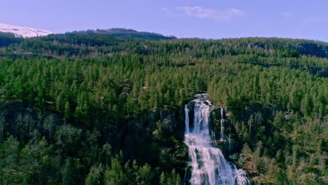 Toma-Aérea-Hacia-Atrás-De-Una-Cascada-Desde-La-Cima-De-Una-Montaña-Rodeada-De-Un-Denso-Bosque-Verde-A-Lo-Largo-De-La-Ladera-De-La-Montaña-Durante-El-Día