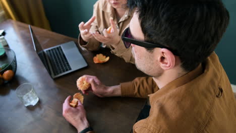 over the shoulder shot of a woman and blind man with glasses eating tangerines and talking while sitting at table at home