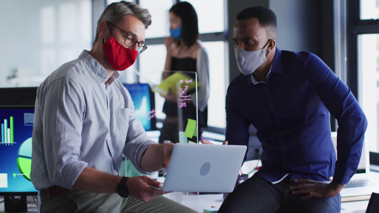 Diverse male office colleagues wearing face masks discussing over laptop at modern office