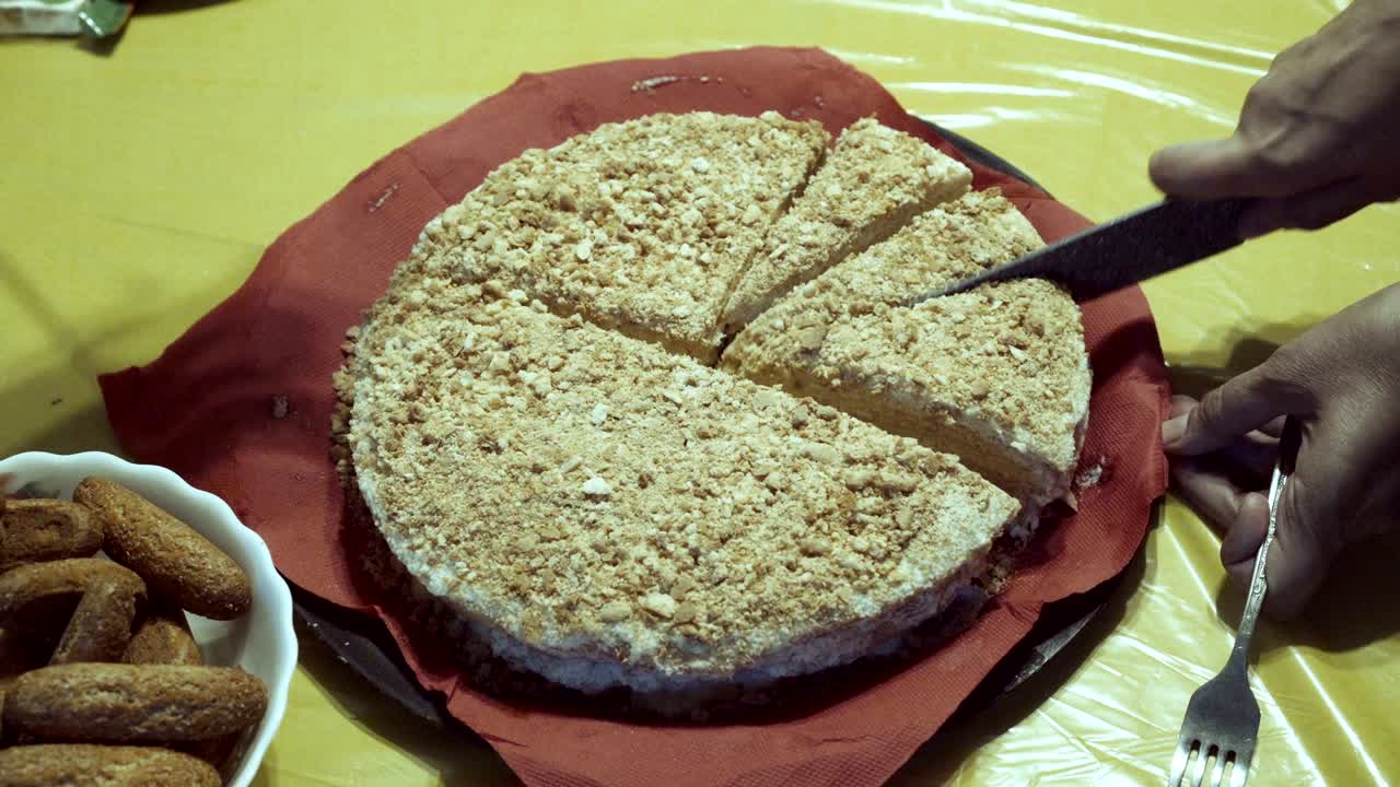 en primer plano, la mamá comienza a cortar el pastel en pedazos con un cuchillo grande y el bebé la ayuda. el pastel está en una servilleta roja sobre un mantel amarillo en la mesa. la vista desde arriba.