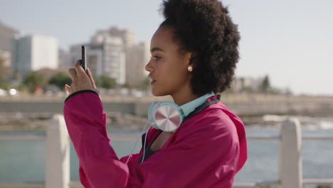 retrato de una atractiva mujer afroamericana en ropa deportiva tomando fotos de la costa usando un teléfono inteligente
