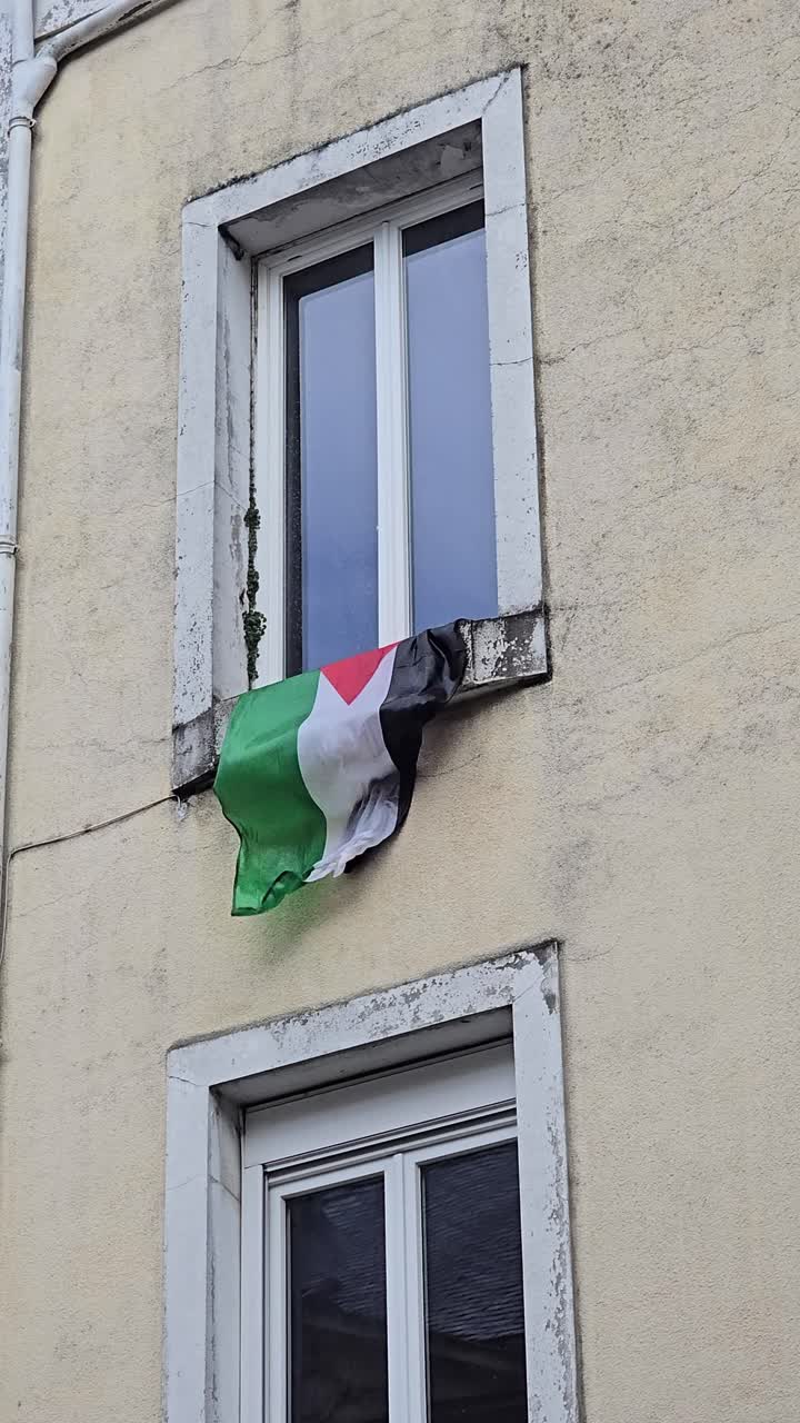 Vertical shot of a Palestinian flag hanging from an apartment window, fluttering strongly in the wind against a beige wall