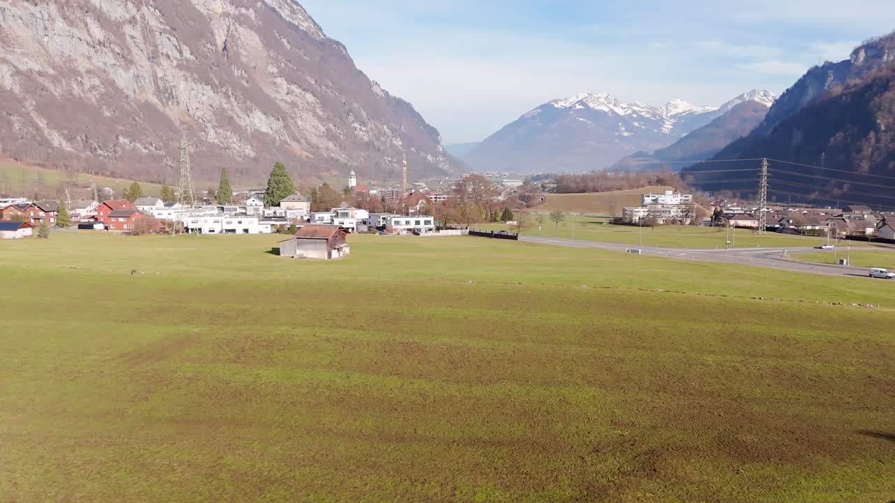 Aerial flyover solar panels on farmstead with green agricultural farm fields in swiss town. Snowy peak of mountain range in Background .Aerial wide shot.
