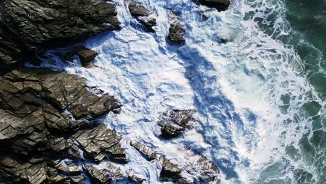 Vista-Giratoria-De-Arriba-Hacia-Abajo-De-Las-Olas-Del-Océano-Y-Aguas-Blancas-Salpicando-Rocas-De-Pizarra-De-Cornualles