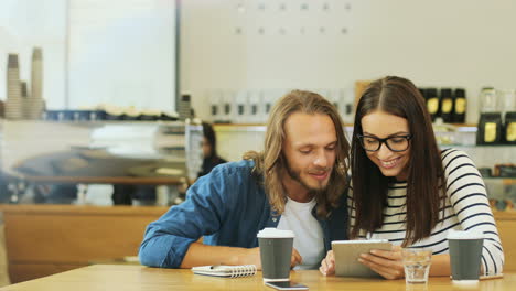 caucasian man and woman friends talking and watching something on a tablet sitting at a table in a cafe