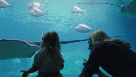 Glückliches-Mädchen-Mit-Mutter-Im-Aquarium-Und-Blick-Auf-Den-Stachelrochen,-Der-Mit-Fischen-Im-Aquarium-Schwimmt.-Kind-Beobachtet-Neugierig-Meerestiere-Und-Hat-Spaß-Daran,-Mit-Mutter-Im-Ozeanarium-Etwas-über-Das-Leben-Im-Meer-Zu-Lernen