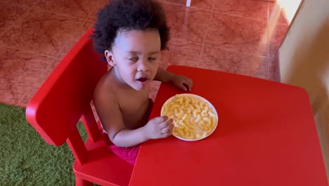 exotic two year old african american kid eating cheese snack seated in a tiny red plastic table