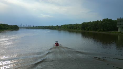 -Red-speedboat-cruising,-distant-sunset-city-skyline,-riverbank-vegetation