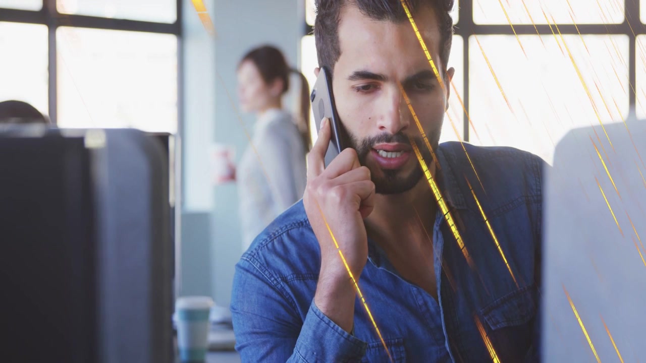 Uomo che risponde a una chiamata in ufficio, tenendo il telefono e guardando un monitor con un overlay giallo, mentre un collega passa dietro.