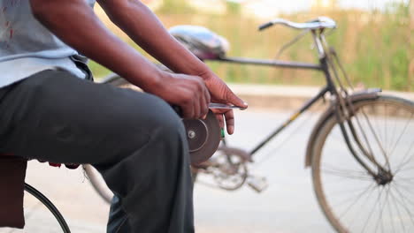 Primer-Plano-De-Las-Manos-De-Un-Hombre-Afilando-Un-Cuchillo-En-Una-Máquina-De-Afilar-Improvisada-De-Bricolaje-En-Un-Marco-De-Bicicleta