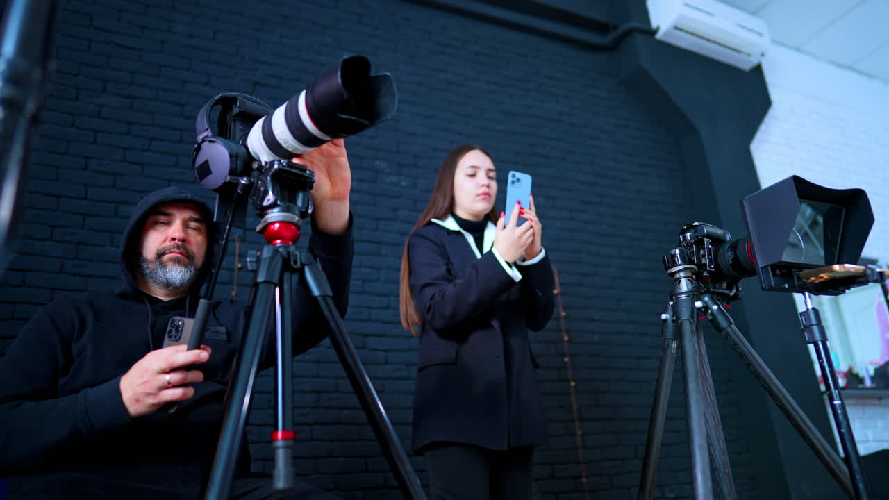 Video štáb připravuje vybavení v tmavé studiové místnosti. Muž a žena nastavují profesionální kamery a chytrý telefon pro natáčení před černou cihlovou zdí