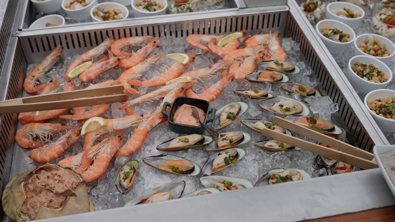 Large buffet tray of whole prawns and open mussels on crushed ice with lemon slices, tongs, dipping sauce, and side dishes visible