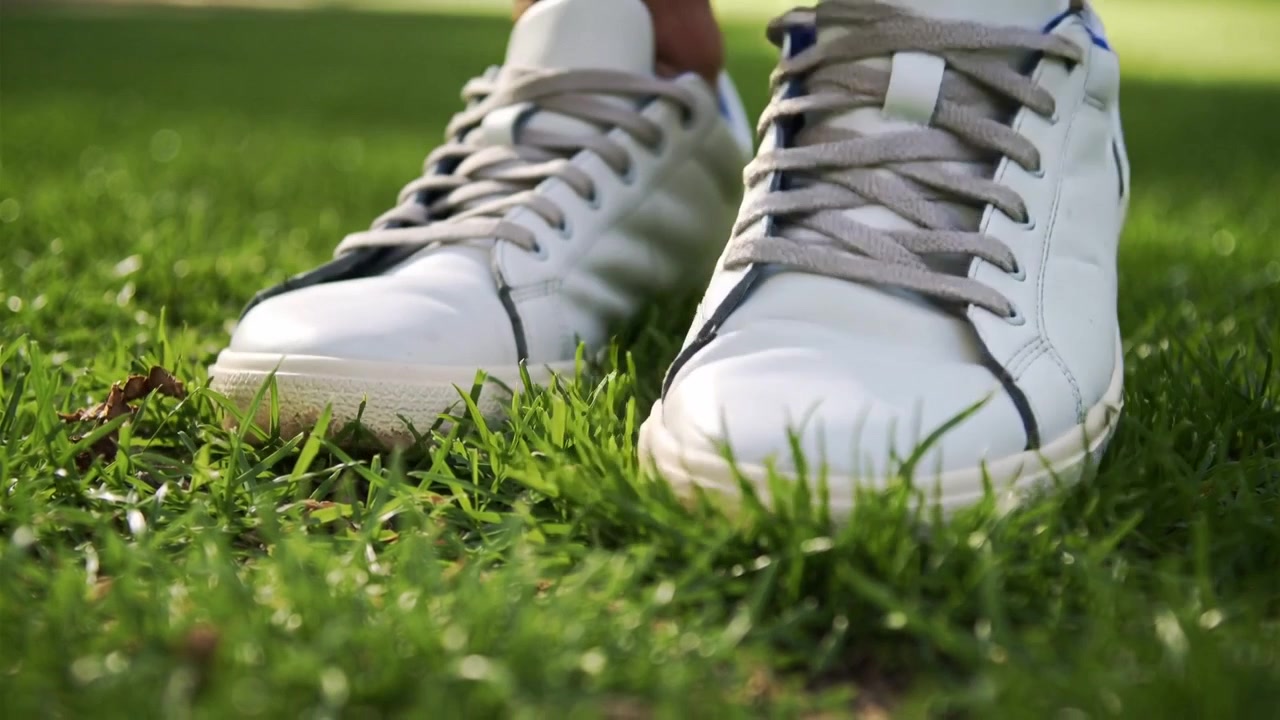 Sollevando una sneaker bassa bianca destra mentre chi la indossa sposta il peso sull'erba illuminata dal sole, mostrando i lacci grigi