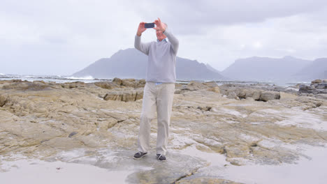 vista frontal de un anciano caucásico haciendo clic en la foto con el teléfono móvil en la playa 4k