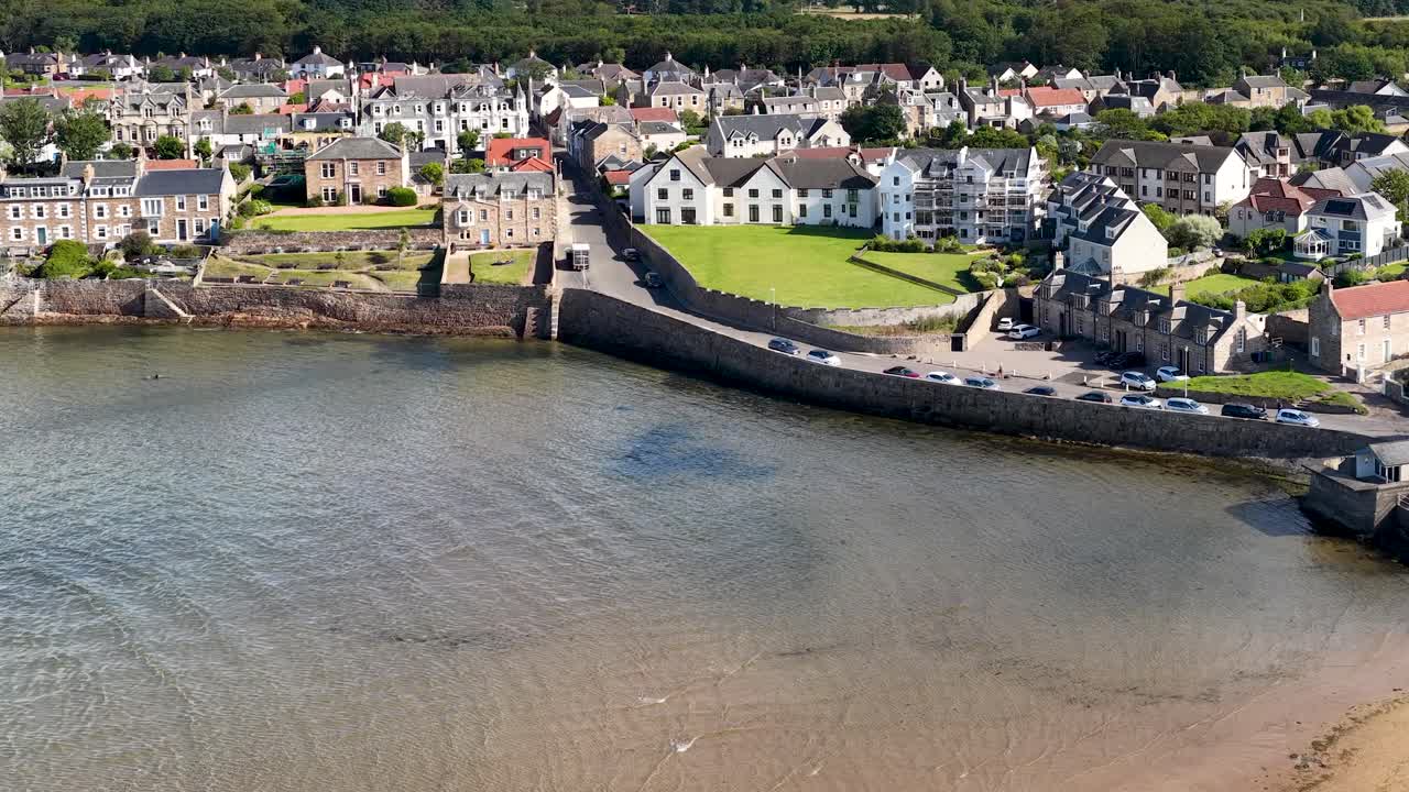 Drone glides toward seaside village, revealing residential houses, seawall, and sandy shoreline in sunlight
