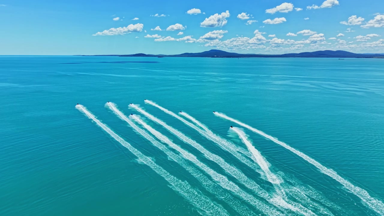 Jetski bevæger sig hurtigt over blåt vand i en udendørs scene, der viser skyer og fjerne bjerge under en klar himmel