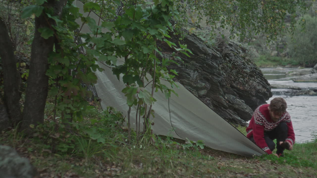 Man Anchoring Tent Edge to Ground near River and Rocks in Forest