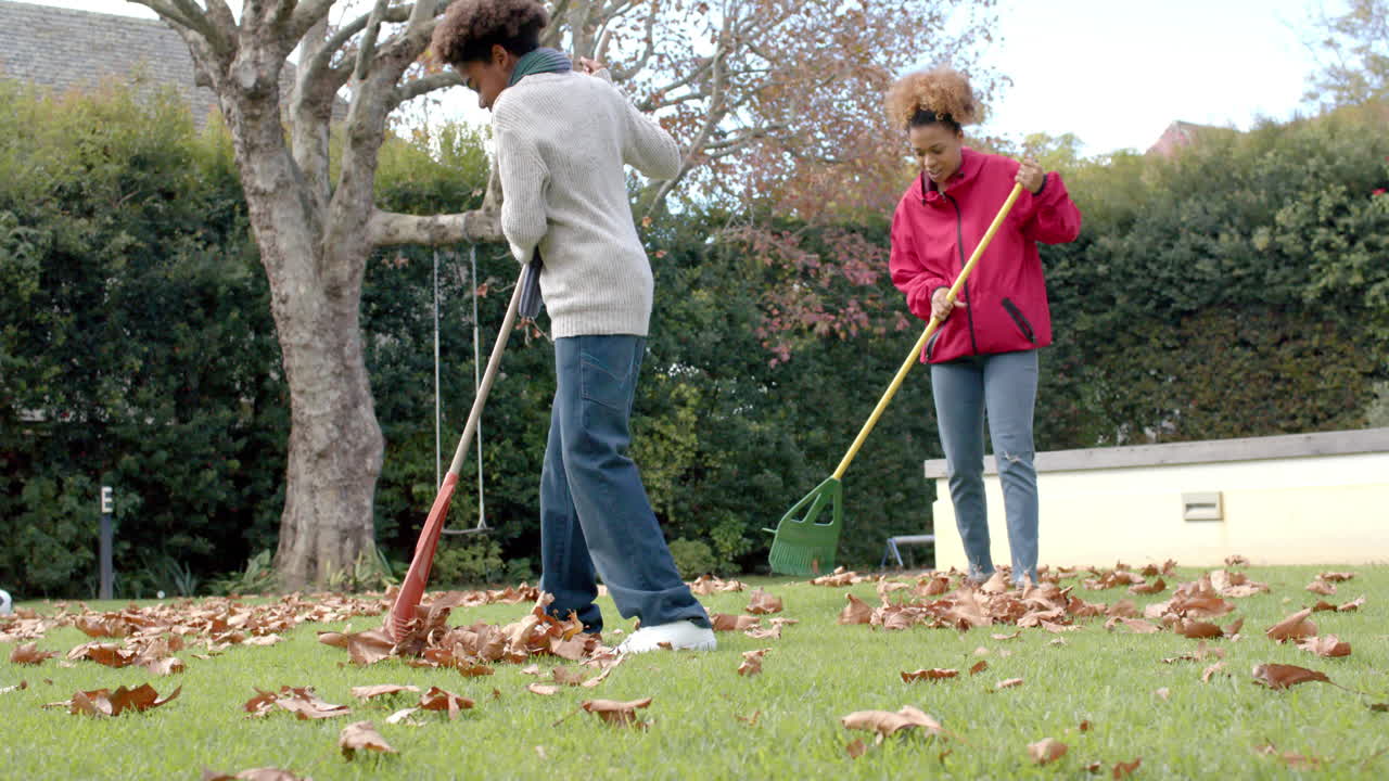 African american mother and son raking up leaves on lawn in garden, in slow motion