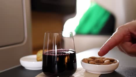 a hand reaches towards a bowl of almonds next to a glass of red wine on a table.