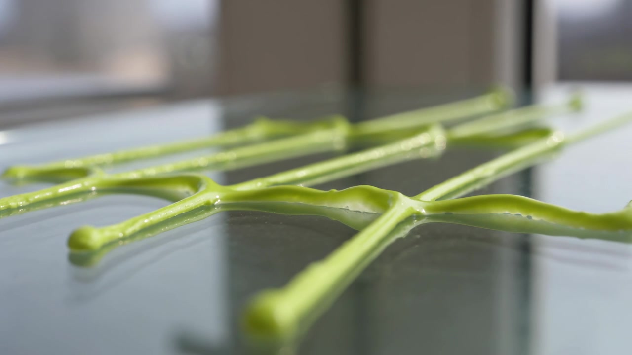 Settling bright green viscous strands on glass near window after extrusion, forming tension ripples