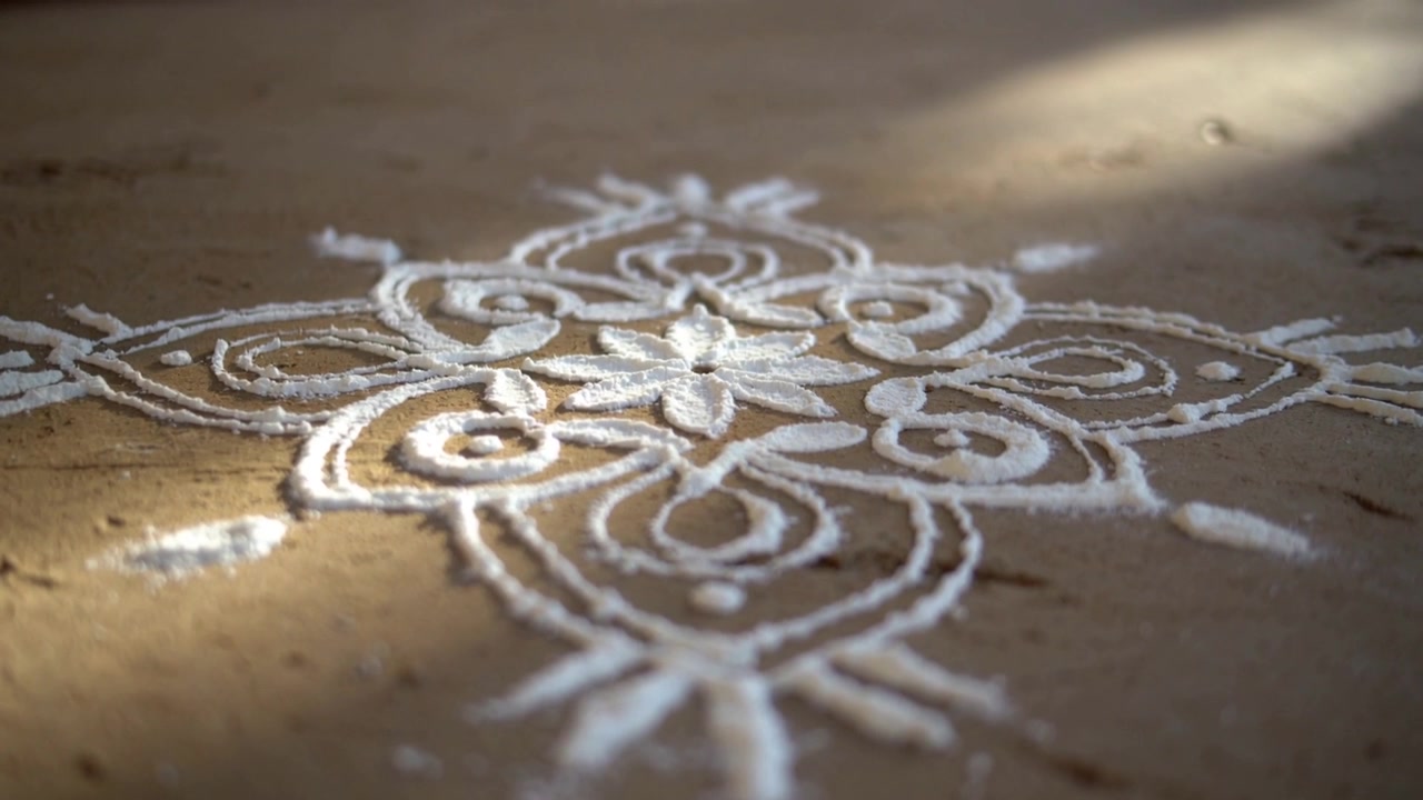 La telecamera si allontana e la messa a fuoco si sposta, rivelando un rangoli bianco sul pavimento della veranda che mostra la sua trama.