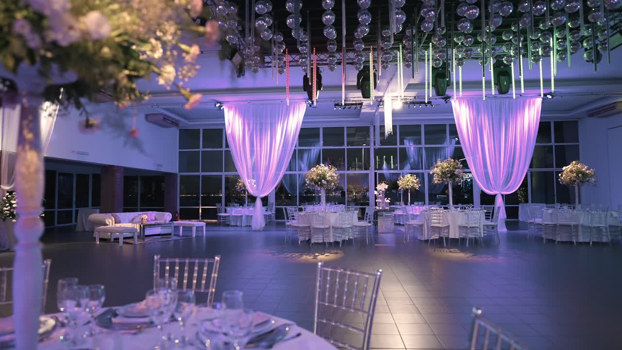 Modern glass‑walled ballroom set for wedding reception, with round tables dressed in white linens, tall floral centerpieces, chiavari chairs, soft purple uplighting on draped curtains and disco balls