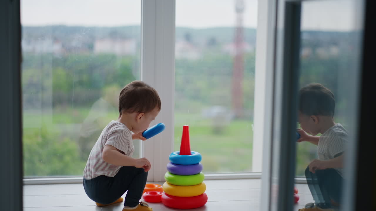 Toddler boy comes up to a colorful pyramid near the panoramic window. Active happy kid assembles the toy.