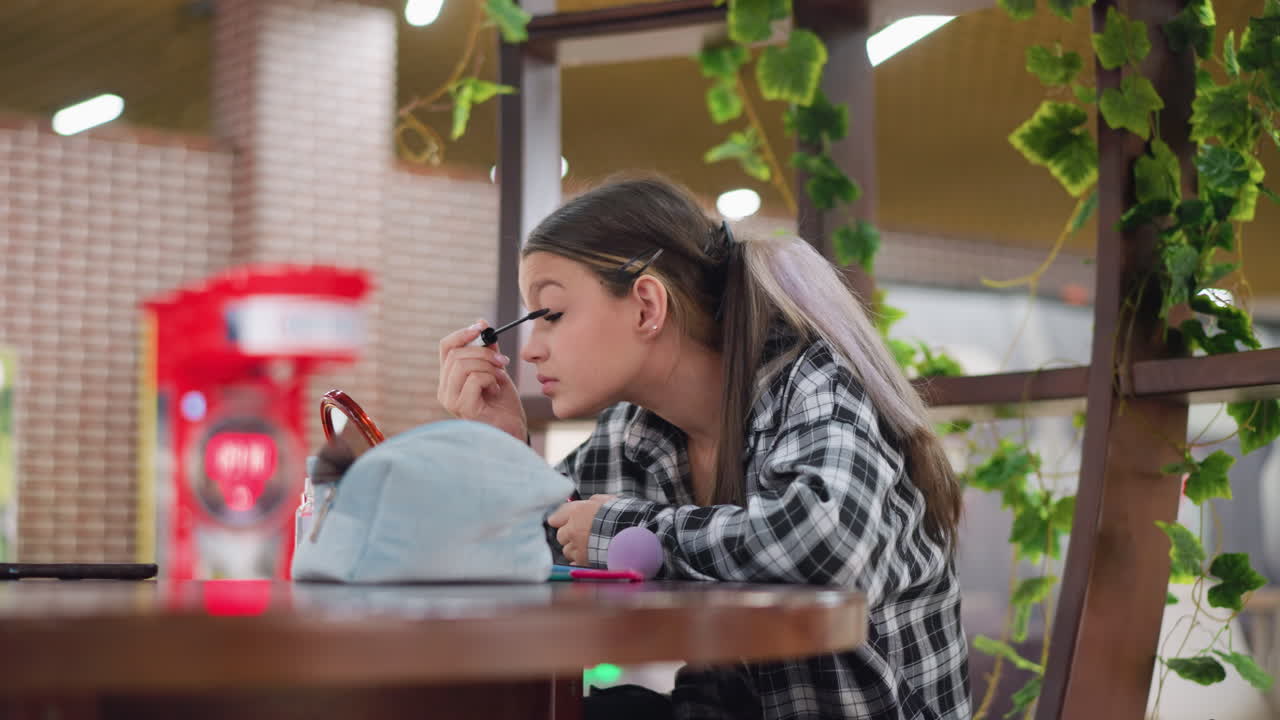 Young lady applying mascara to her eyelashes while sitting at a table with makeup products in front, focusing on eyelash grooming with a blurred background of plants and a vibrant public space