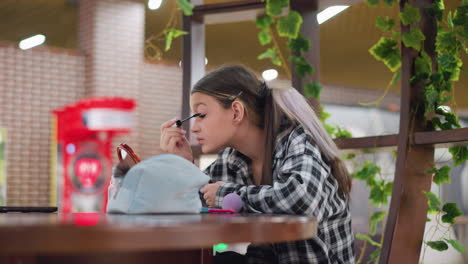 una joven que aplica máscara a sus pestañas mientras está sentada en una mesa con productos de maquillaje al frente, centrándose en el cuidado de las pestañas con un fondo borroso de plantas y un espacio público vibrante
