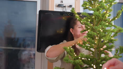 Close-up of woman in beige gown decorating Christmas tree with lights, arranging branches with care, family preparing for holiday celebrations with Christmas tree decorations