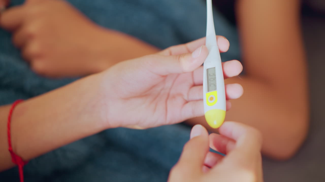 Close up of hands holding thermometer with clear digital display reading 36 degrees Celsius while someone lies down blurred in background showing monitoring care and health check