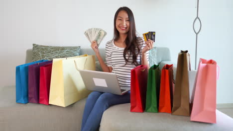 satisfied and happy looking attractive woman sitting an a couch wearing a striped shirt and blue jeans smiling with money in one hand and credit cards in the other surrounded by colorful shopping bags