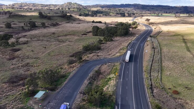 Two large trucks travel along a winding rural highway in open countryside, captured by a smoothly moving aerial camera in natural daylight