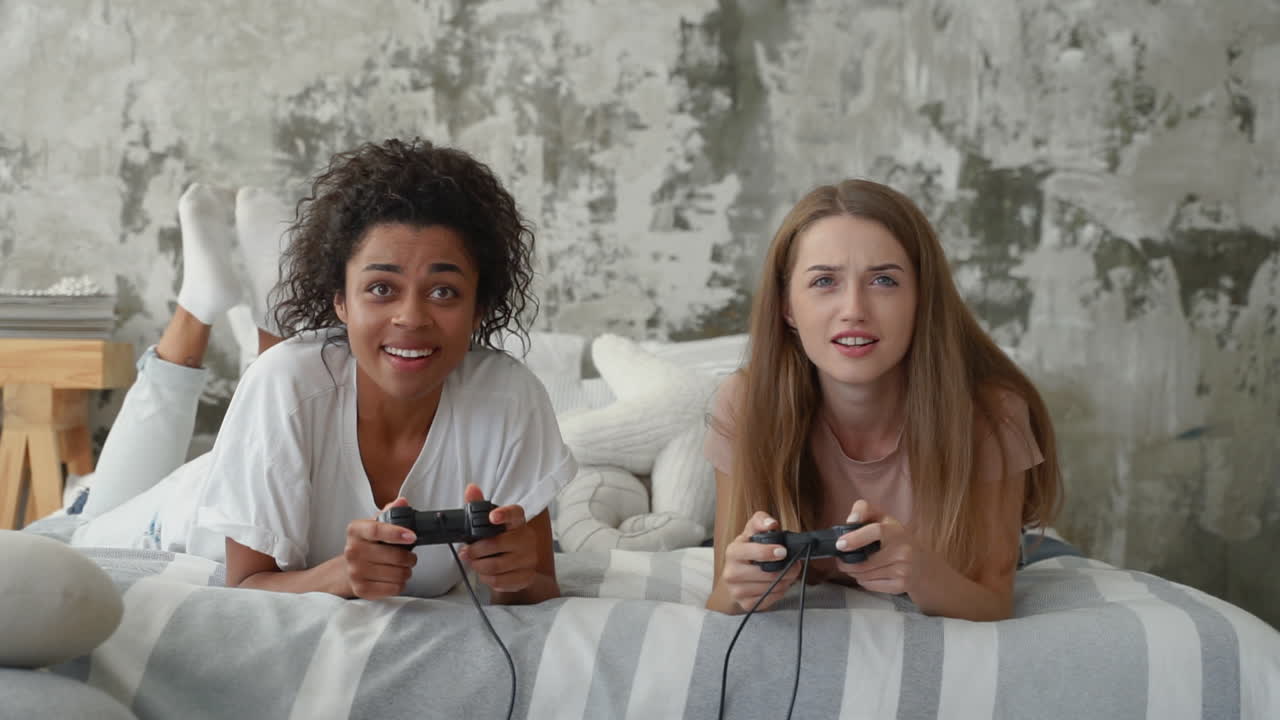 dos amigas jugando videojuegos. una chica negra y una mujer joven caucásica sosteniendo controladores y divirtiéndose acostados en la cama.