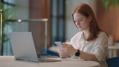 Red-haired-Woman-working-from-home-using-laptop-computer-while-reading-text-message-on-mobile-phone.-Woman-using-a-phone.-Serious-charming-woman-using-smartphone-while-working-with-laptop-at-home