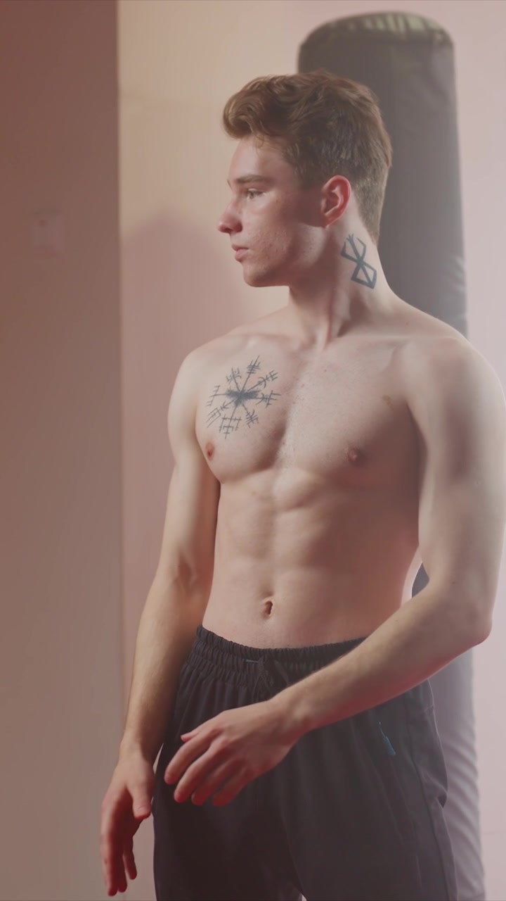 Giovane uomo con tatuaggi solleva la maglietta nera per rivelarli. I tatuaggi includono simboli runici sul petto vicino a forme geometriche. La maglietta viene rimossa gradualmente dall'alto verso il basso esponendo più corpo.
