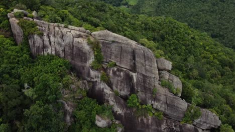 Aerial-shot-of-a-woman-sitting-on-top-of-a-large-cliff-looking-over-the-forest,-majestic-untouched-nature